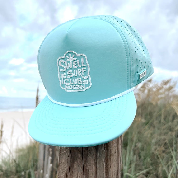 Mint green cap with 'Swell Surf Club' logo on a wooden post with beach background
