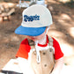 Child wearing a cap with 'Noggin' text, playing with a small drum outdoors.