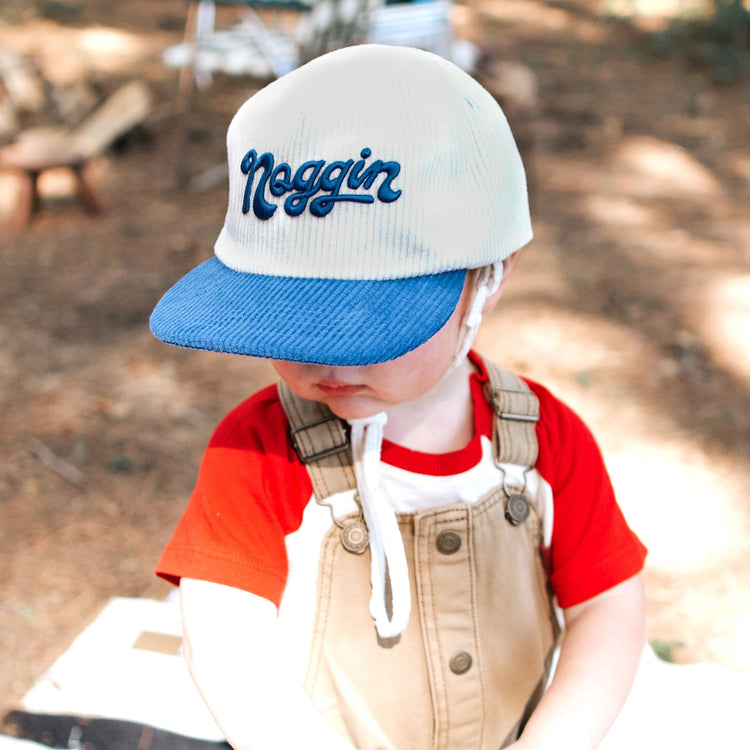 Child wearing a cap with 'Noggin' text, playing with a small drum outdoors.