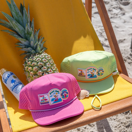Two colorful caps with pineapple and beach-themed stickers on a yellow beach chair with a pineapple and sunscreen tube.