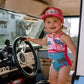Child in a colorful swimsuit and pink cap standing inside an old-fashioned vehicle.