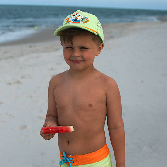 Young boy on a beach holding a watermelon slice, wearing a cap and swim shorts.
